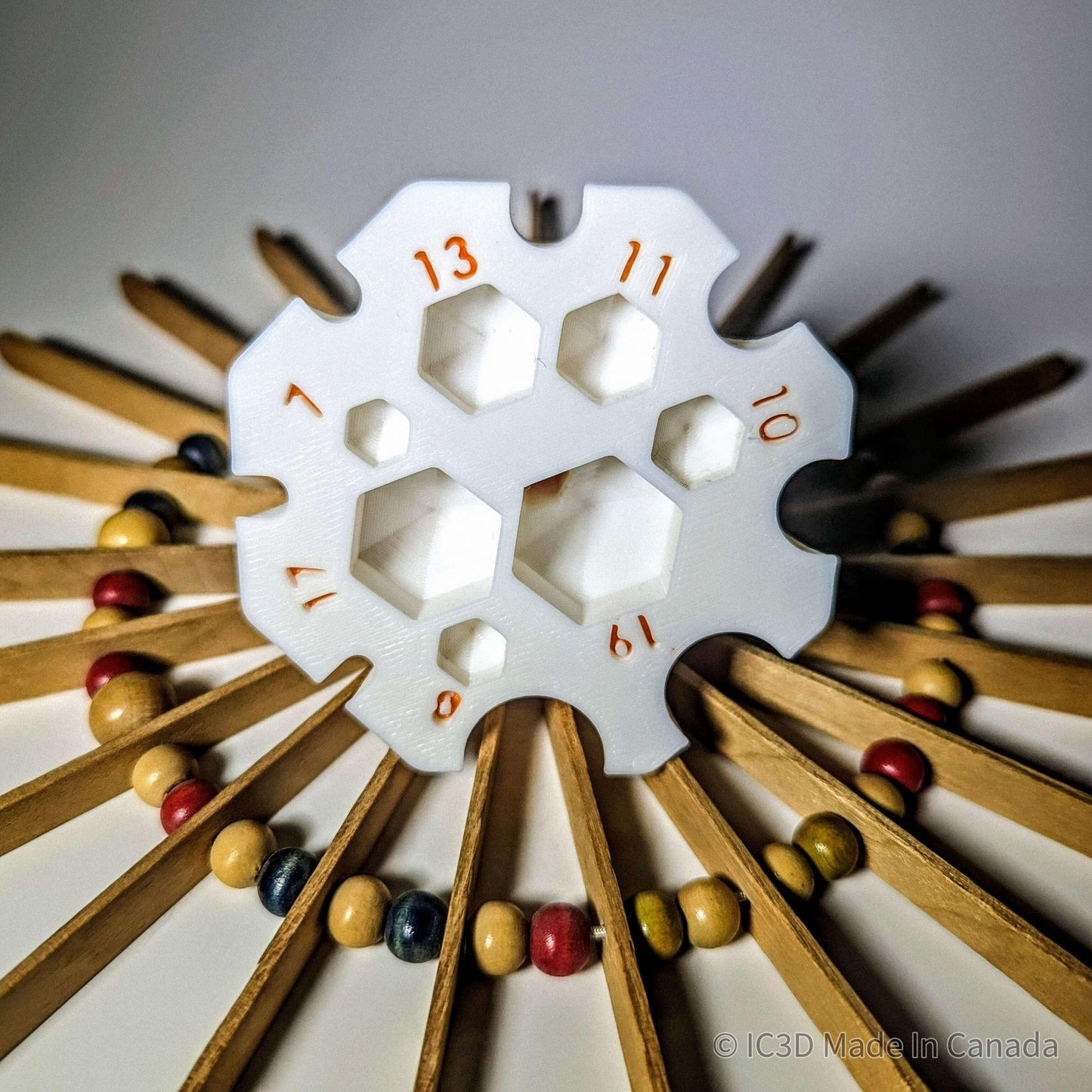 3D printed white yarn gauge tool with hexagonal holes, surrounded by wood beads and sticks